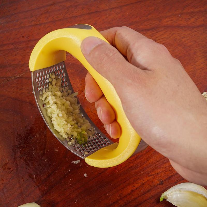 Hand using yellow handled stainless steel garlic press to mince garlic on wooden surface