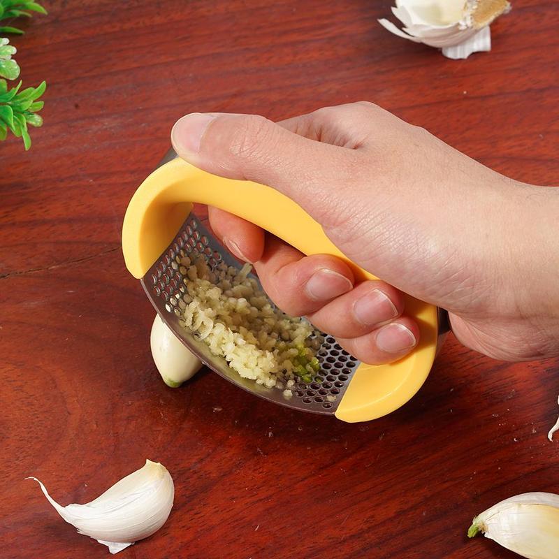 Hand using yellow handle stainless steel garlic press to crush garlic cloves on wooden surface