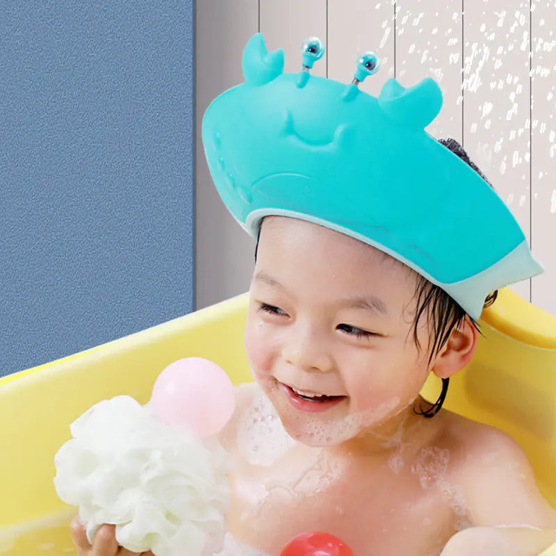 Happy toddler wearing a blue adjustable baby shower cap shaped like a crab while playing in the bath