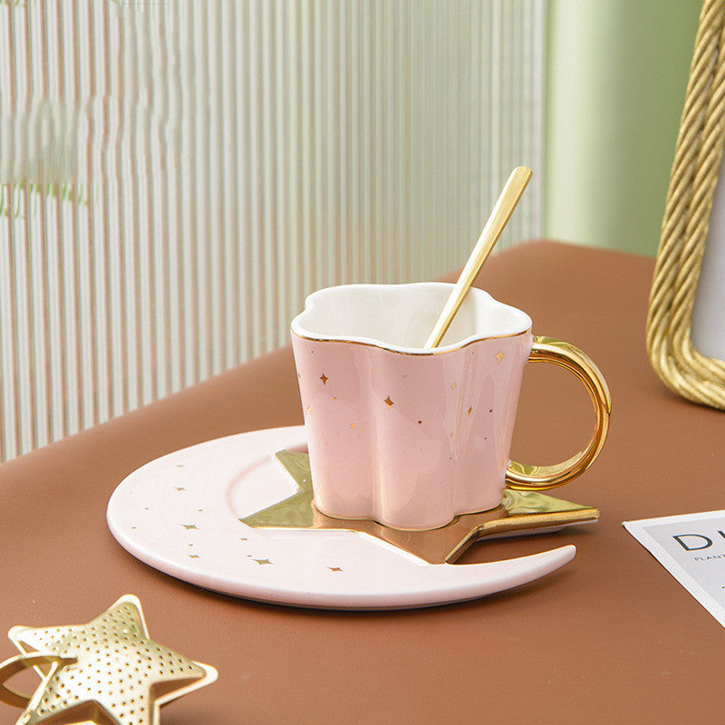 Pink ceramic cup with star and moon saucer featuring gold handle and accents on a brown table