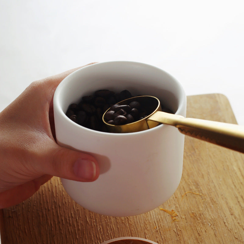 Hand holding a white ceramic kitchen storage jar with a gold scoop filled with coffee beans