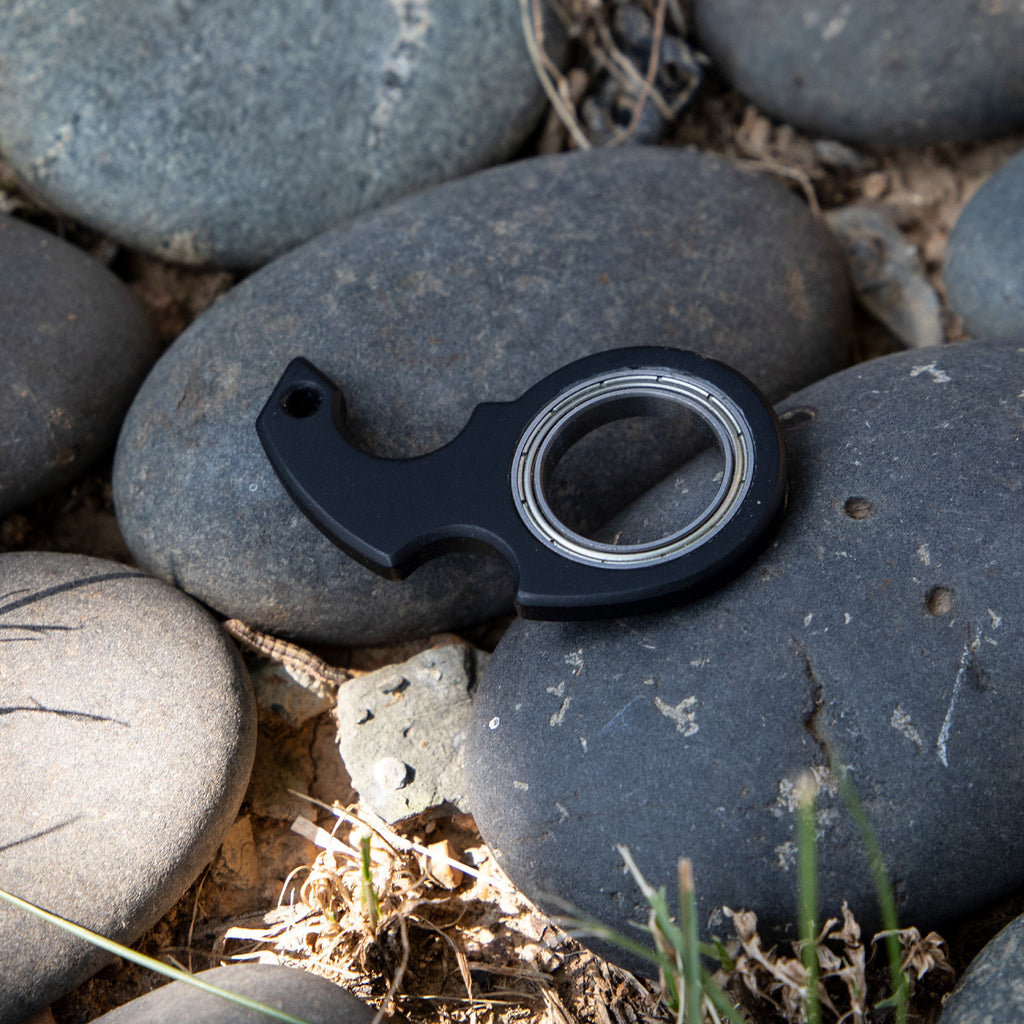 Shopi Vella black finger spinner bottle opener resting on smooth gray stones outdoors