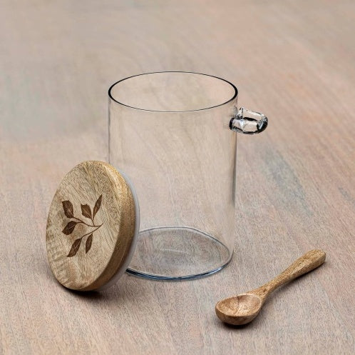 Shopi Vella glass kitchen storage jar with wooden lid and carved leaf design beside wooden spoon