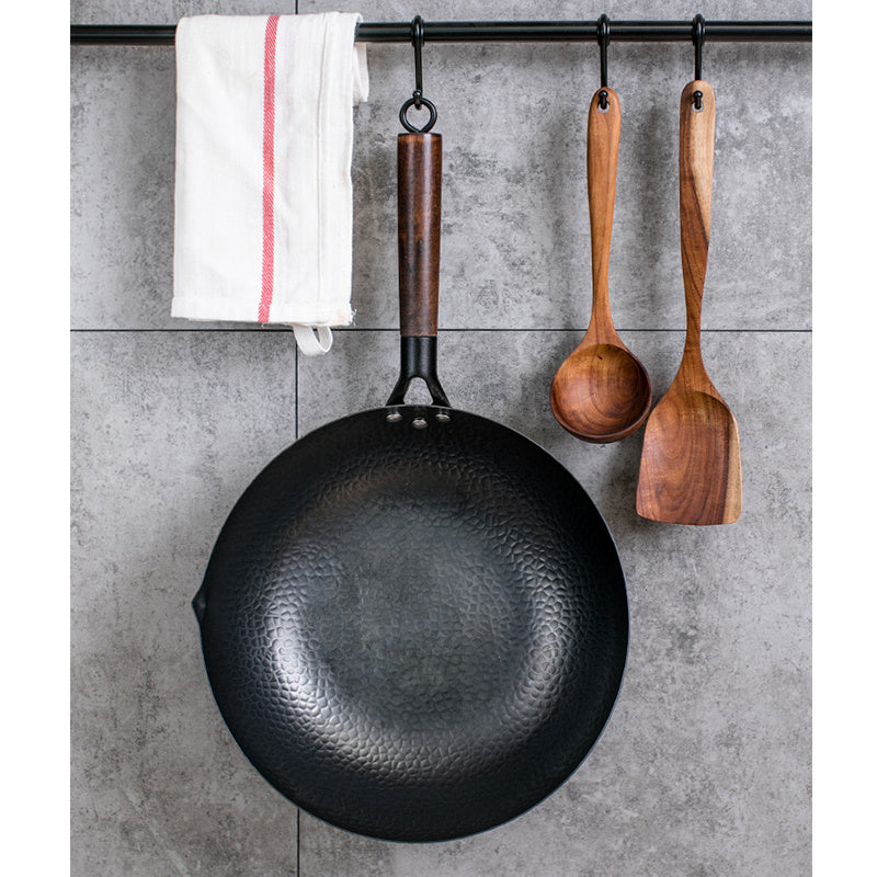 Handmade iron wok with wooden handle hanging beside wooden cooking utensils and a kitchen towel