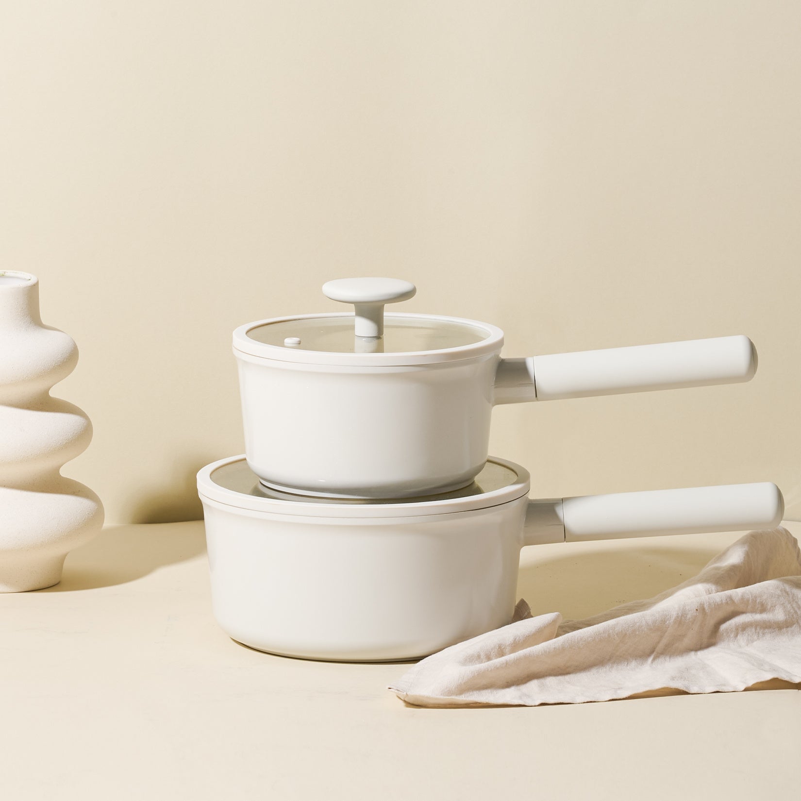 White healthy induction cookware with removable handles stacked on beige surface with cloth and decorative vase