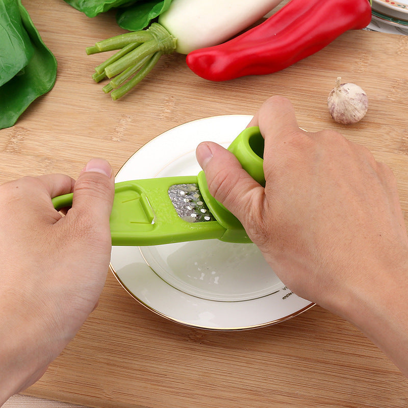 Hands using green garlic masher kitchen tool to press garlic over a white plate on wooden surface
