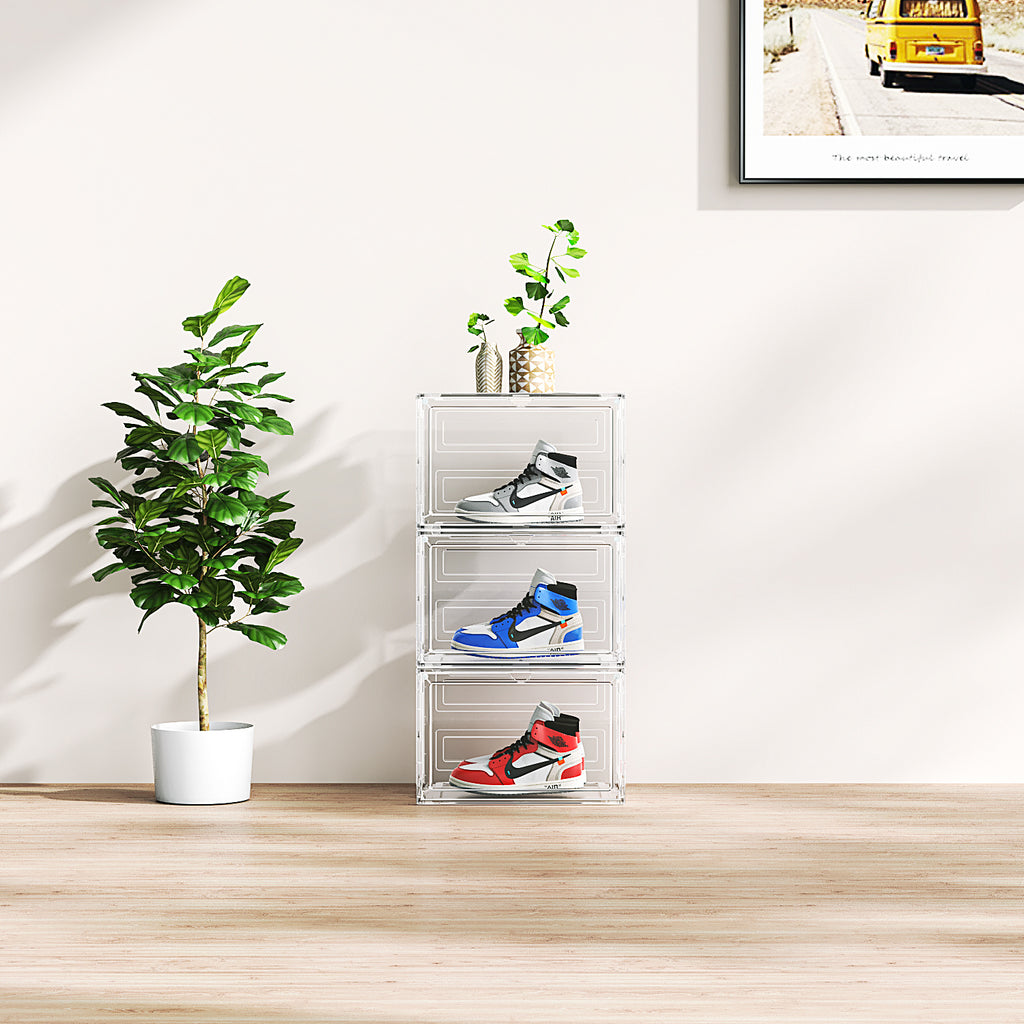 Stacked large acrylic shoebox organizer with sneakers inside next to a potted plant in a bright room