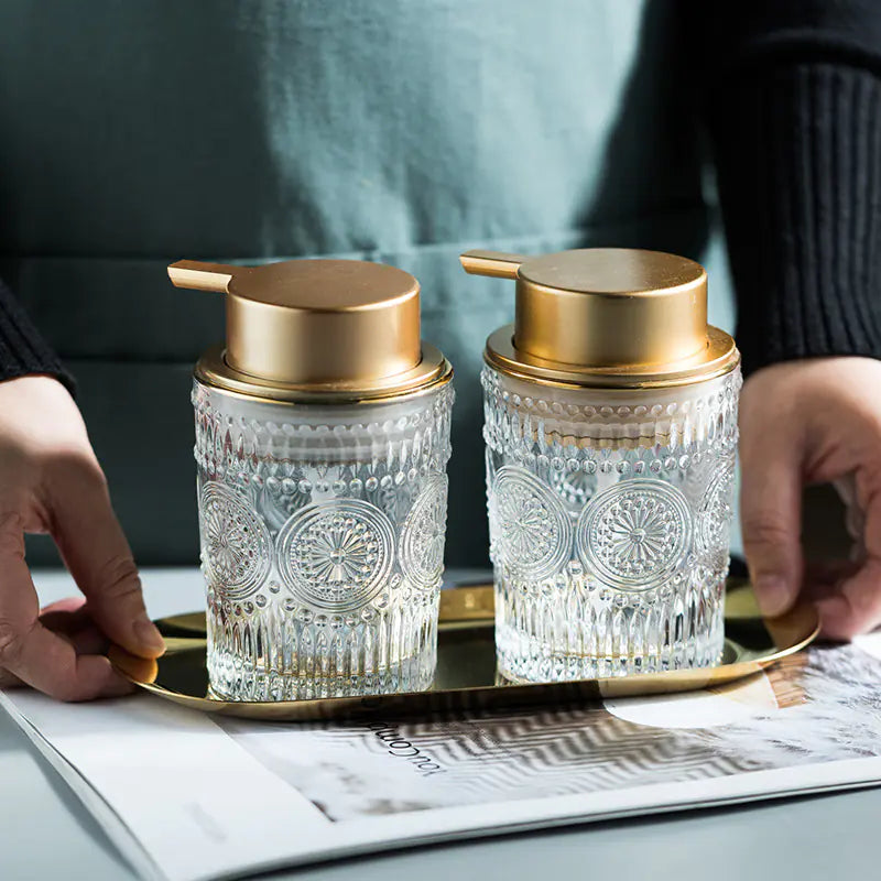 Luxury glass soap dispenser set with gold pump tops on a gold tray held by a person