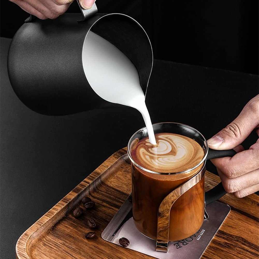 Black stainless steel milk frothing pitcher pouring milk into coffee on wooden tray