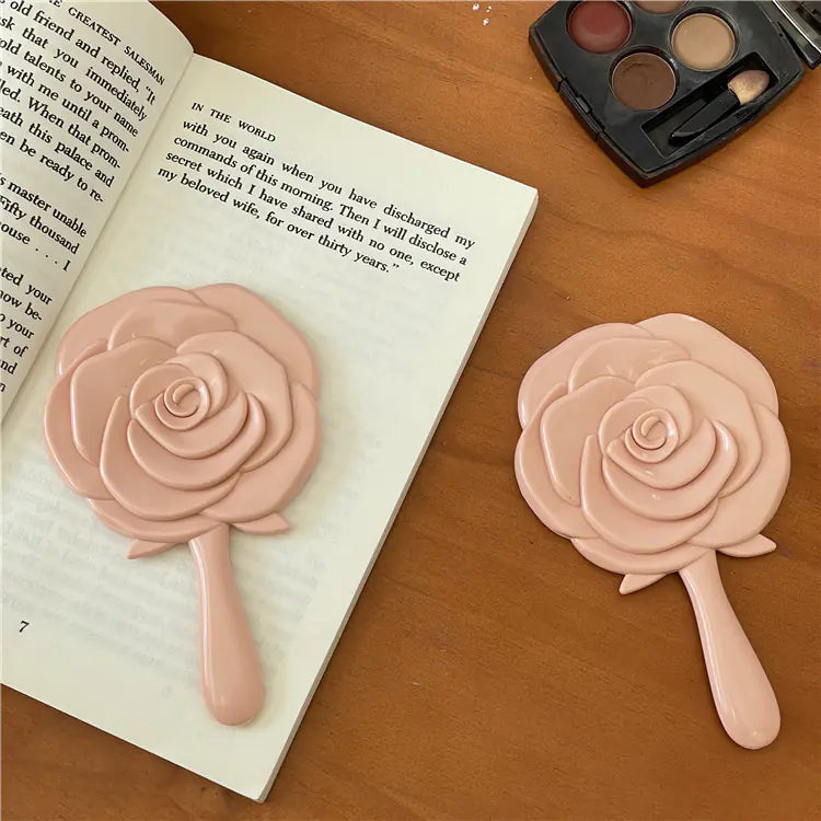 Pink rose shaped portable cosmetic mirror for students with handle on wooden surface and open book