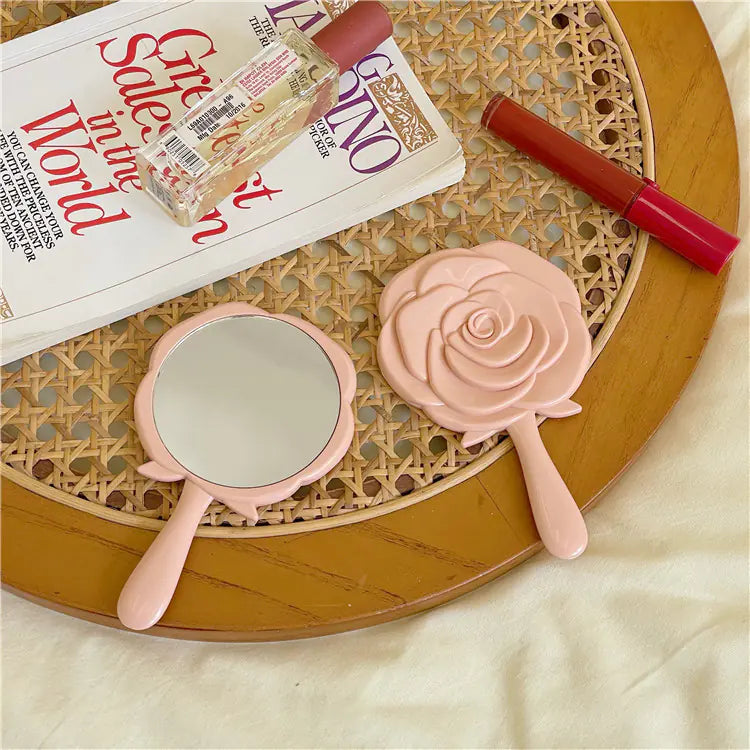 Pink portable cosmetic mirror for students with rose design on wooden table with magazine and lipstick