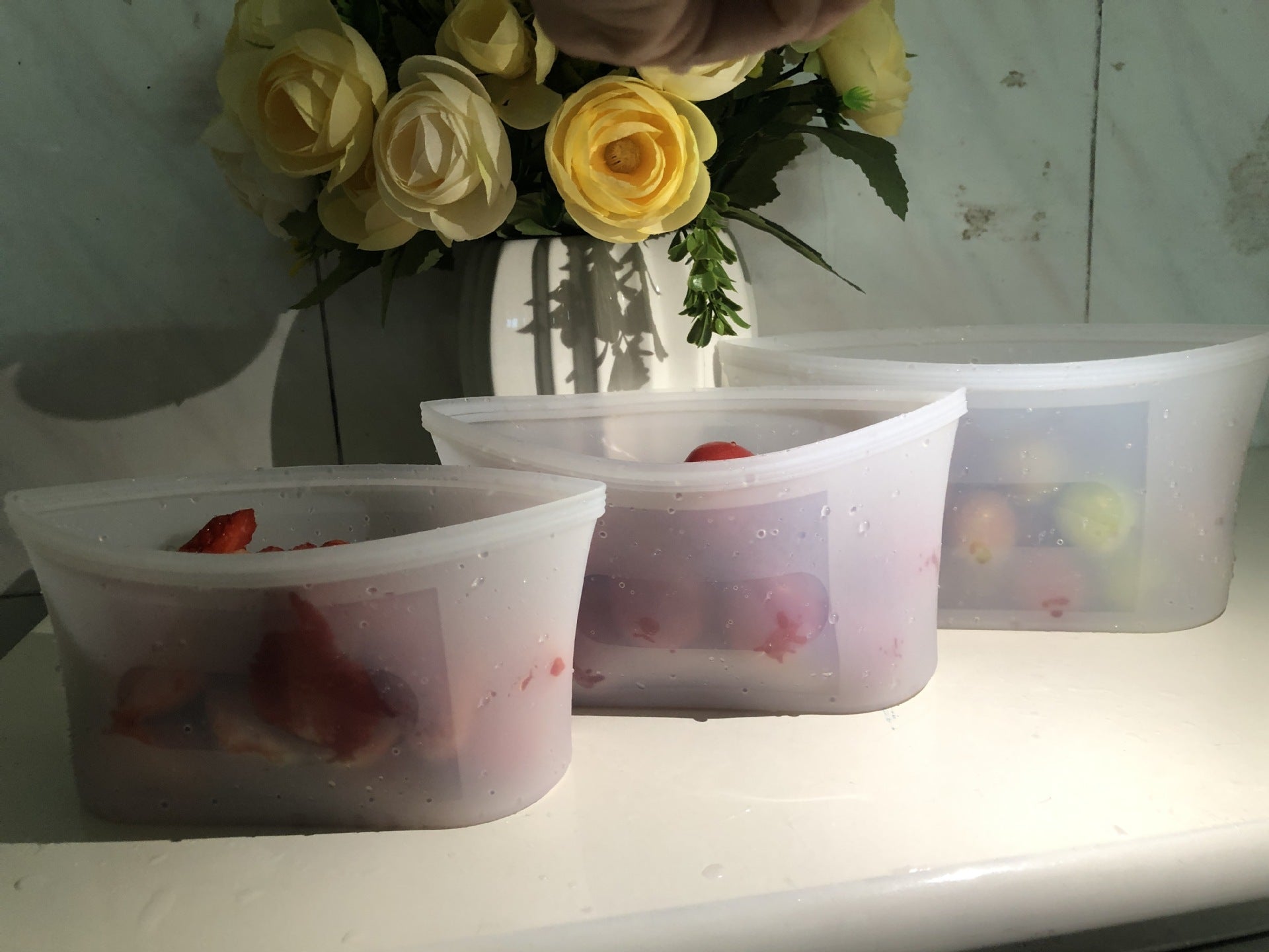 Shopi Vella reusable silicone food storage bags holding fruits on a white surface with yellow roses in the background