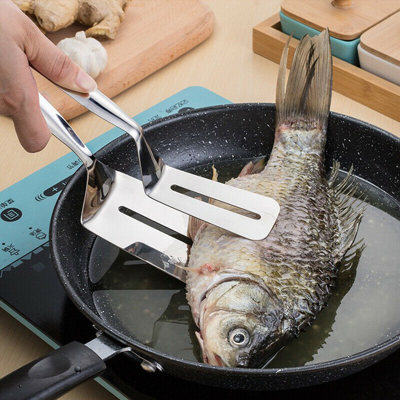 Stainless steel kitchen tongs flipping a whole fish in a black frying pan on a stovetop