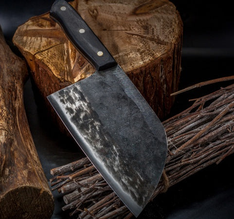 Shopi Vella stainless steel meat cleaver with black handle resting on rustic wood logs