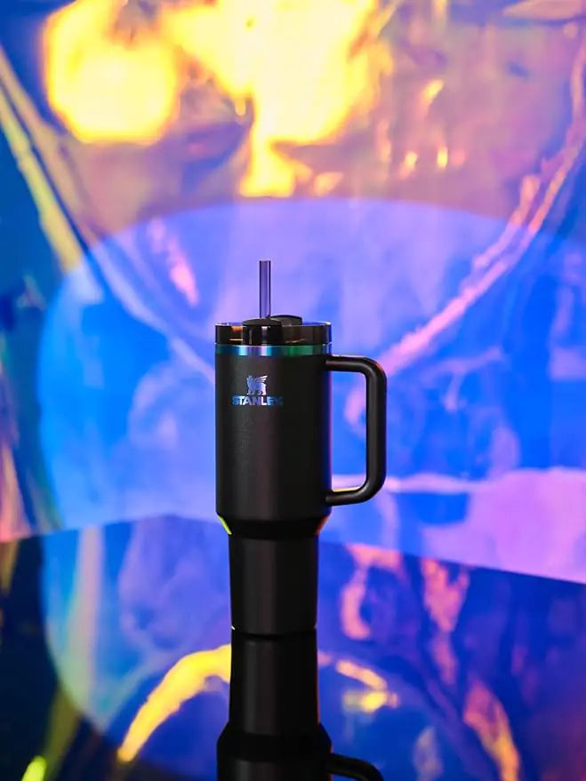 Black Stanley tumbler with straw and handle placed on reflective surface with colorful background