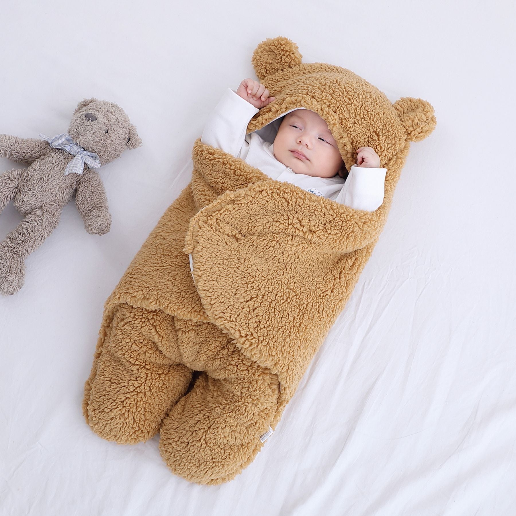 Baby wrapped in a thick winter baby quilt with bear ears lying on a white bed next to a teddy bear