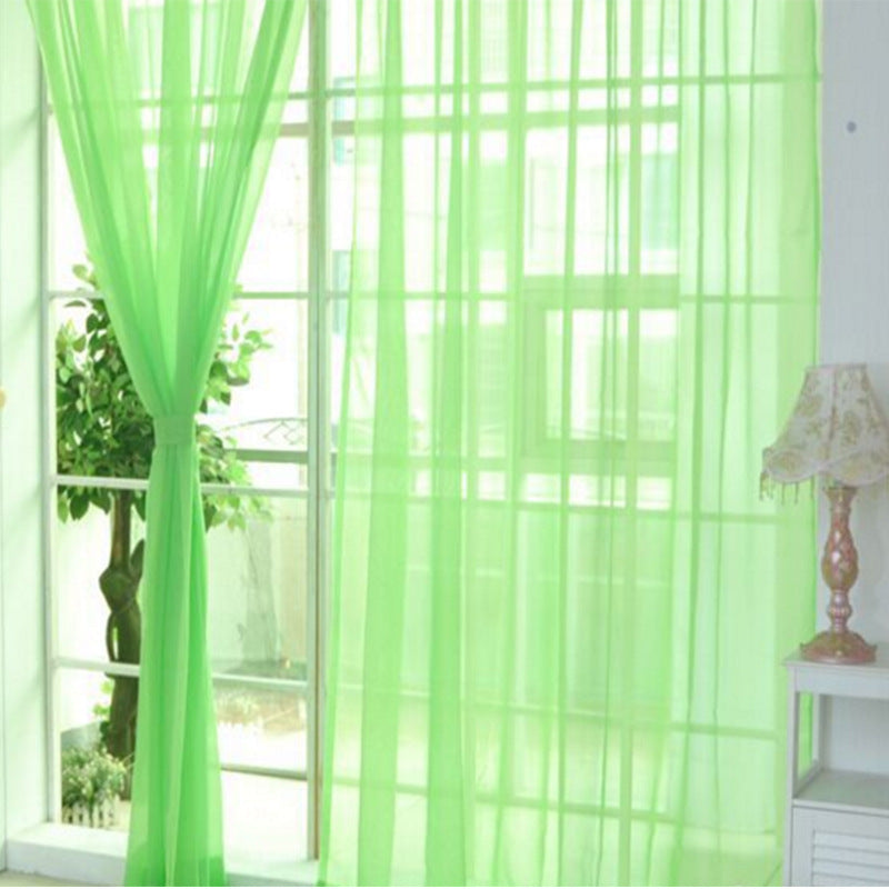 Green transparent tulle window curtains hanging by a bright window with a lamp and plant nearby