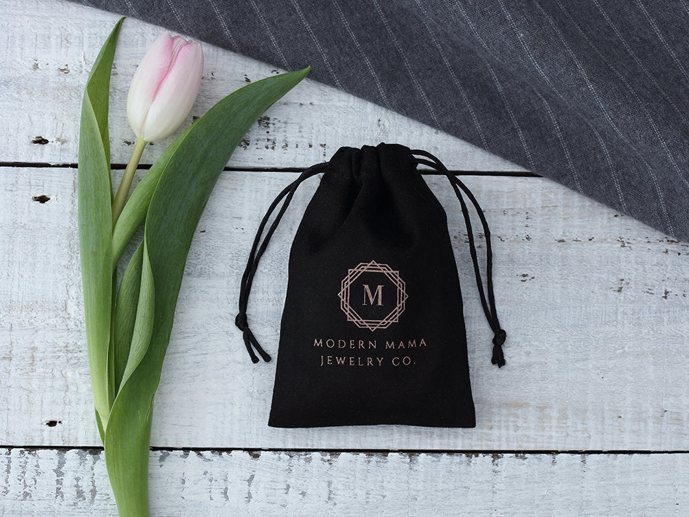 Black velvet jewelry gift bag with Modern Mama Jewelry Co logo next to a pink tulip on white wooden surface