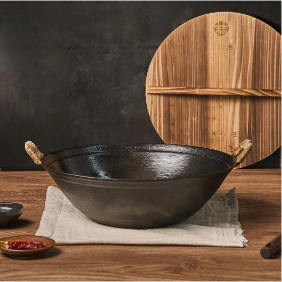 Handmade cast iron wok pan with rope handles on wooden table and wooden lid in background