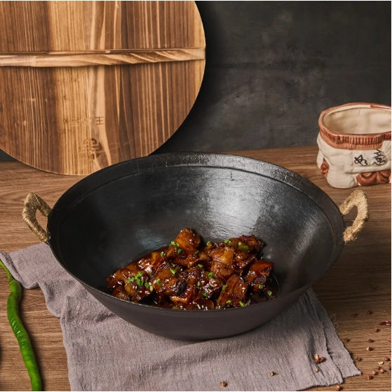 Handmade cast iron wok pan with cooked meat dish on wooden table with green chili and rustic decor