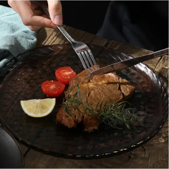 Stainless steel cutlery set used to cut a cooked steak on a black glass plate with tomatoes and lemon wedge
