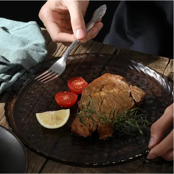 Person holding fork from stainless steel cutlery set eating steak with cherry tomatoes and lemon on black glass plate