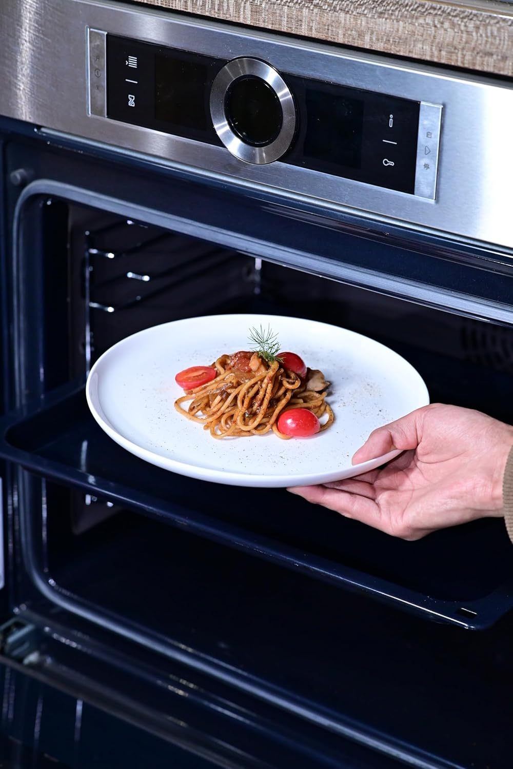 Hand placing white dinnerware set for 4 plate with pasta and tomatoes into modern stainless steel oven
