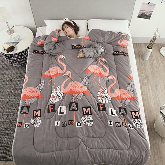 Gray winter quilt with sleeves featuring pink flamingo design by Shopi Vella on bed with woman wearing sleep mask