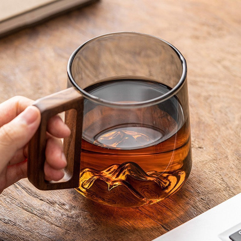 Hand holding wooden handle glass cup filled with amber liquid on wooden surface